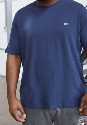 Mand iført en marineblå kortærmet skjorte og grå shorts med et lille logo-patch på brystet af skjorten og benet på shortsene.