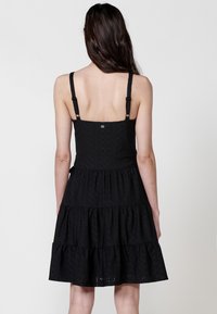 Schwarzes, schichtetes Kleid mit verstellbaren Trägern, das aus strukturiertem Stoff mit filigranen Mustern besteht. Das Design hat ein tailliertes Oberteil und einen ausgestellten Rock.