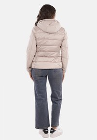 Giacca puffer cappuccio di un beige chiaro con sezioni trapuntate orizzontali, abbinata a jeans blu e sneakers bianche, vista da dietro.