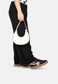 Schwarze Sneakers aus strukturiertem Material, mit weißen Akzenten und einer braunen Gummisohle. Eine weiße, halbrunde Handtasche wird in einer Hand gehalten.