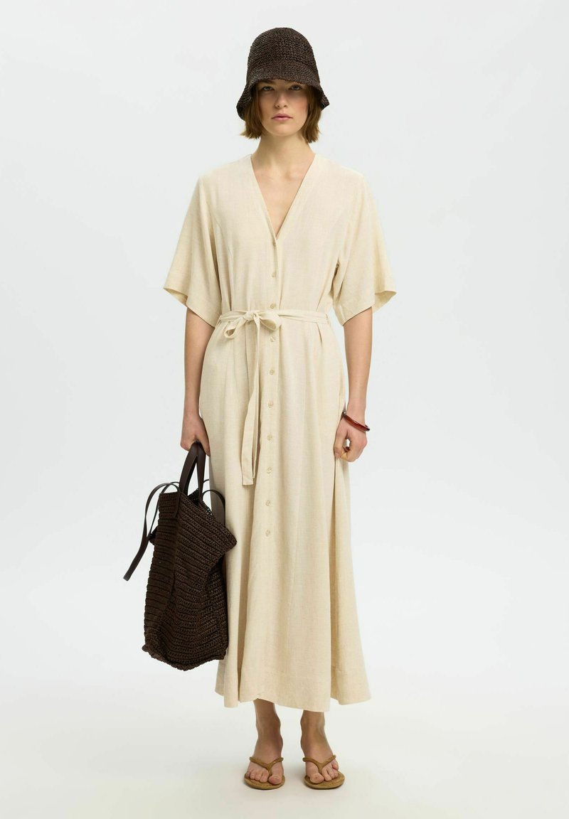 Femme portant une robe beige boutonnée avec ceinture à la taille, chapeau bob crochet marron, tenant un grand sac tressé marron et portant des sandales beige.