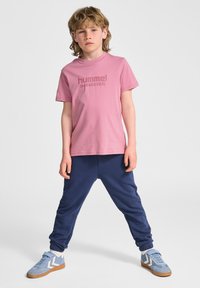 Lyserød bomulds t-shirt med prægede "hummel SPORTSWEAR" logo, parret med marinefarvede joggers og lysblå ruskinds sneakers med brune såler.