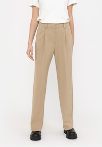 Beige skräddarsydda byxor med rak passform, framfållar och en slät textur, tillsammans med en enfärgad vit t-shirt och kraftiga svarta skor.