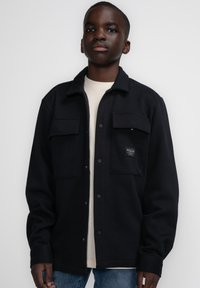 Zwarte wolmix jas met drukknoopsluiting, twee borstzakken en een label. Draag het met een crèmekleurig shirt en blauwe spijkerbroek.