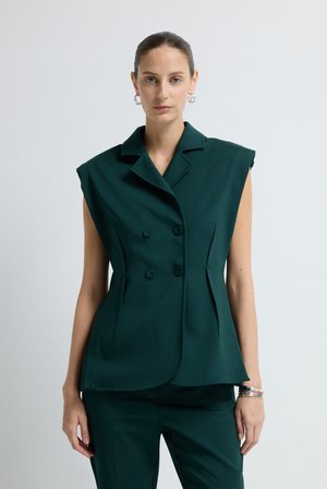 Vrouw met strak teruggebonden haar, gekleed in een donkergroene mouwloze dubbele blazer en bijpassende broek, staand tegen een effen witte achtergrond.