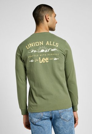 Maglietta verde a maniche lunghe con una stampa grafica sul retro che presenta il testo "UNION ALLS" e "realizzata con uno scopo" in crema.