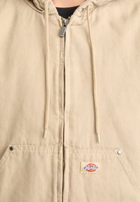 Khaki hooded jacket with front zipper, drawstrings, and two lower pockets. Features silver hardware and a logo patch on the left pocket.