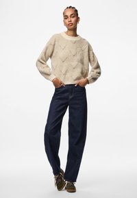 Mujer de pie con las manos en los bolsillos, vestida con un suéter beige con patrones, jeans oscuros y zapatillas verdes, frente a un fondo blanco.