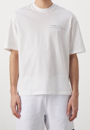 Biały T-shirt z krótkim rękawem z małym czarnym tekstem "Armani Exchange" na górnej lewej części klatki piersiowej, noszony przez osobę z rozluźnionymi ramionami.