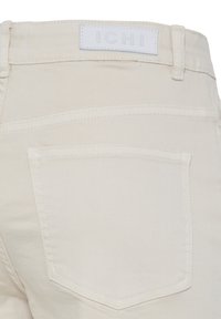 Hele hele beežist denimpiidest teksade tagakülg, kus on nähtav tagatasku ja valge silt vöökohal kaubamärgiga "ICHI".