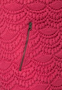Gros plan d’un tissu rose avec des motifs complexes de dentelle circulaire et une fermeture éclair noire verticale au centre.