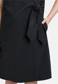 Schwarzes Kleid aus glattem Stoff mit seitlicher Bindung und Tasche. Gerader Schnitt mit abgerundetem Saum am Boden.