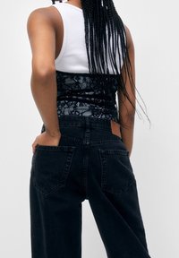 Jeans de mezclilla negros de cintura alta con piernas anchas, con un forro de encaje texturizado y bolsillos traseros divididos. Camiseta de tirantes blanca por debajo.