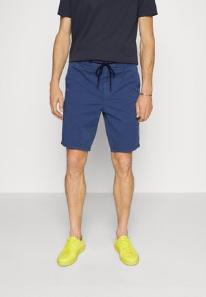 Man met marineblauwe korte broek met een zwart trekkoord, een zwart t-shirt en felgele sneakers, staand tegen een effen achtergrond.