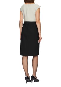 Robe noire avec une silhouette ajustée, manches courtes en crème et fermeture éclair au dos. Jambes visibles avec des collants transparents et des talons hauts noirs.