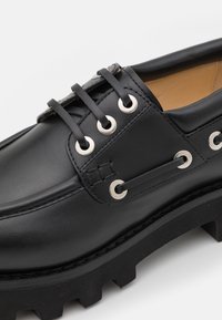 Chaussure en cuir noire avec semelle épaisse à crampons, lacets noirs passés dans des œillets argentés, et intérieur de col rembourré beige clair.
