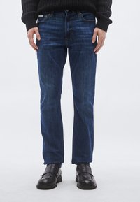 Vaqueros de mezclilla azul oscuro con ajuste entallado, que cuentan con bolsillos estándar, detalles sutiles de costura y efecto desgastado, combinados con mocasines negros.