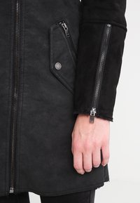 Musta vetoketjullinen takki, valmistettu pehmeästä kankaasta, jossa on pystykaulus, sivutaskut ja teksturoitu pinta hopeisilla metalliyksityiskohdilla.