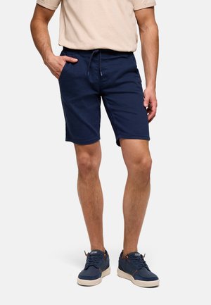 Hombre con camisa de manga corta beige, pantalones cortos azul marino con cordón y zapatillas azul marino, de pie con una mano en el bolsillo.