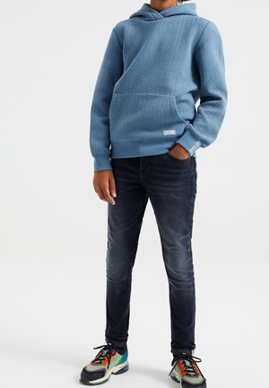 Niño con sudadera azul con capucha y estampado en zigzag, jeans oscuros y zapatillas multicolores, de pie con una mano en el bolsillo.