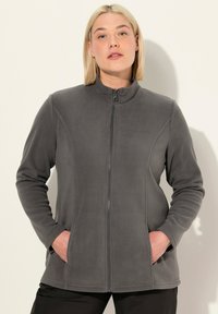 Femme blonde portant une veste en polaire grise à fermeture éclair, les mains dans les poches, se tenant contre un fond clair et uni.