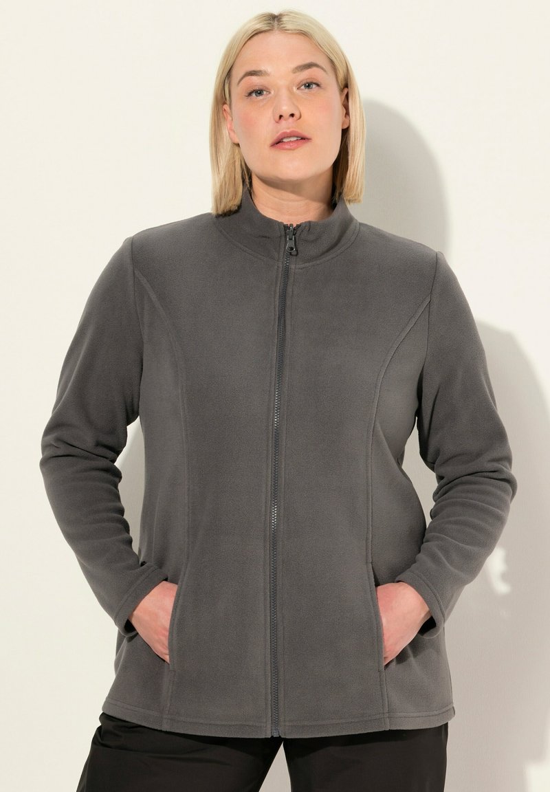 Blond kvinna som bär en grå zip-up fleecejacka med händerna i fickorna, stående mot en plain ljus bakgrund.