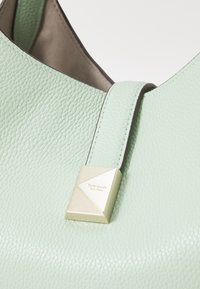 Light green textured leather handbag with a smooth, triangular gold-tone accent featuring engraved brand logo. Simple and modern design.