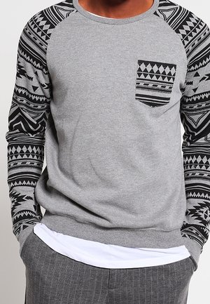 Sweat-shirt gris avec des motifs géométriques noirs sur les manches et la poche poitrine, porté sur une chemise blanche avec un pantalon à fines rayures grises.