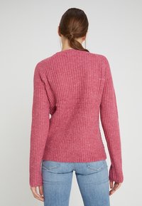 Ribbed, long-sleeve sweater in a dusty pink color, featuring a slightly fitted cut and textured fabric, paired with light blue jeans.