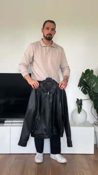 Giacca in pelle nera con zip frontale, colletto e tasche laterali, esposta su un appendiabiti. Il modello indossa una maglietta chiara e pantaloni scuri.