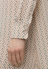 Main d’adulte portant une chemise beige clair avec un motif répétitif de petites raquettes de tennis bleues et de balles de baseball marron.