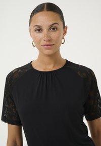 Melnā īsā piedurkņu blūze ar mežģīņu elementiem uz piedurknēm, savāktu kaklu un mīkstu, gludu auduma tekstūru. Vienkāršs, klasiskā dizains.