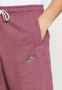 Ruka osoby v kapse bordových Nike tepláků s bílou šňůrkou a třpytivým Nike logem na levém stehně.