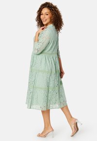 Abito in pizzo verde menta con maniche a tre quarti, vita stretta e gonna ampia. Sandali con tacco a blocco trasparente completano il look.