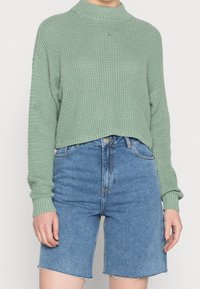 Ljust grönt stickat tröja med en hög, ribbad hals, kombinerat med blå denimshorts med fransad nederkant och klassisk femficksdesign.