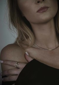 Auksinės spalvos kaklo grandinėlė su mažomis kvadratinėmis jungtimis, sluoksniuotos žiedų su tekstūruotais akcentais, dėvint juodą viršų, demonstruojanti lygią odą ir lakuotus nagus.