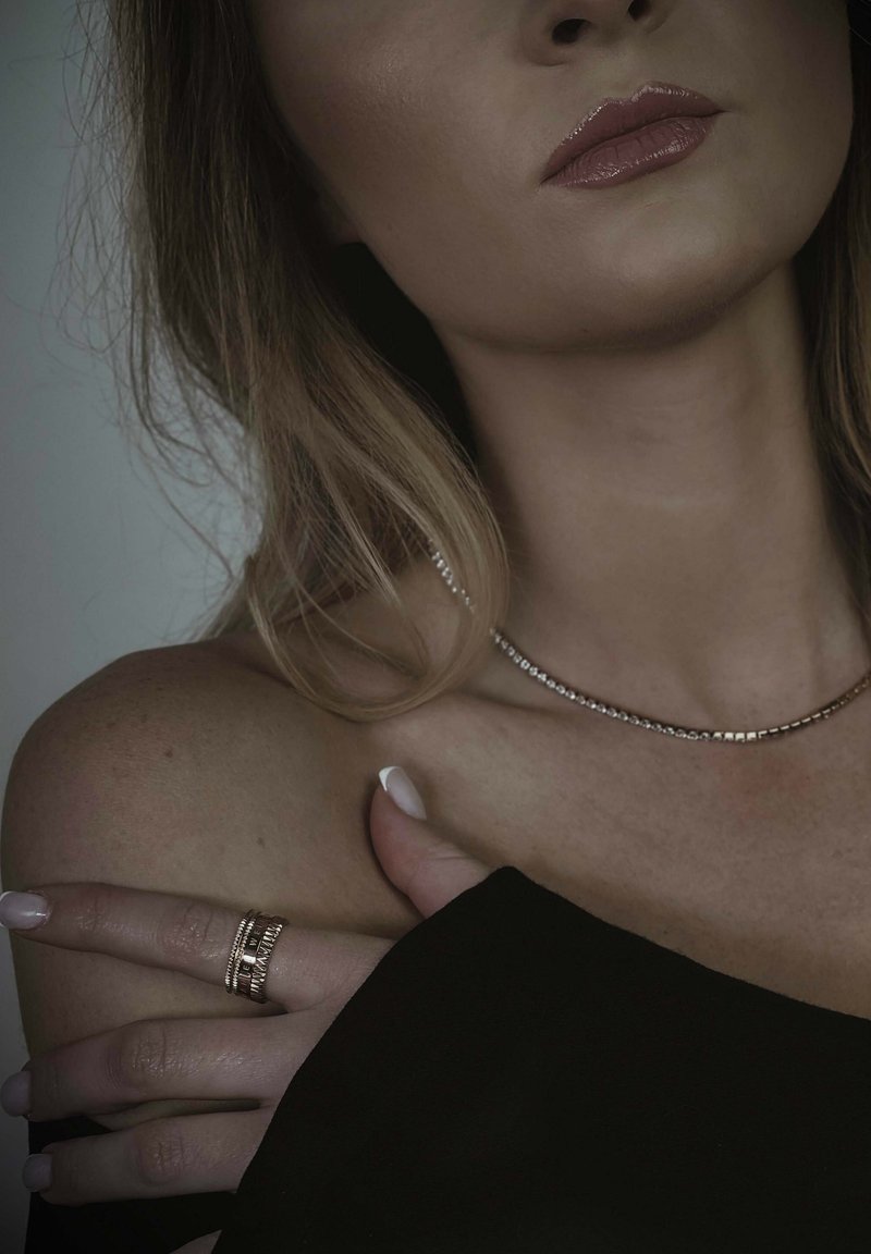 Auksinės spalvos kaklo grandinėlė su mažomis kvadratinėmis jungtimis, sluoksniuotos žiedų su tekstūruotais akcentais, dėvint juodą viršų, demonstruojanti lygią odą ir lakuotus nagus.