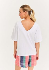 Žena s blond culíkem, oblečená v bílé halence s véčkovým výstřihem na zádech a v mnohobarevných pruhovaných šortkách, stojící na neutrálním světlém pozadí.