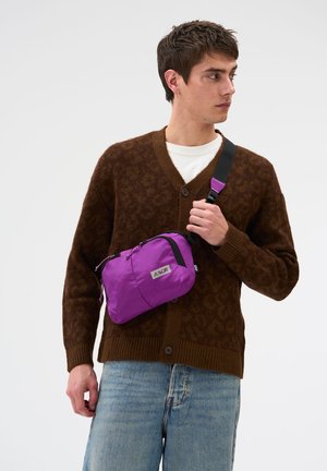 Young man wearing a brown cardigan and light blue jeans holding a purple crossbody bag with a black strap across his chest.