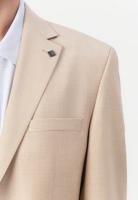 Beige Blazer aus glattem Material mit einer einzelnen Brusttasche und einem kleinen schwarzen Metall-Detail am Kragen. Darunter ein hellblaues Hemd.
