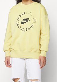 Žlutá bavlněná mikina s černým logem Nike a textovým designem, pružné manžety a lem, pohodlný střih.