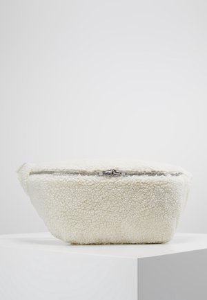 Banane en polaire sherpa blanche avec fermeture éclair double argentée, présentée sur un socle blanc sur un fond clair uni.