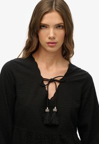 Blusa negra texturizada con escote en pico y cierre de lazo. Presenta borlas con acentos de cuentas. Mangas largas y bajo acampanado.