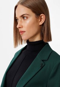 Green blazer with a structured collar, worn over a black turtleneck. Features a small black emblem on the lapel. Smooth fabric texture.