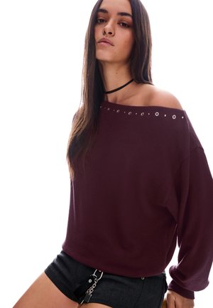 Pull en marron à épaules dénudées avec des œillets en métal argenté le long du col, associé à un short en denim noir avec des accents en métal.
