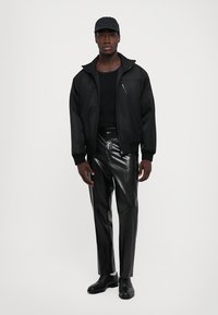 Man som står klädd i en helt svart outfit inklusive keps, zip-up jacka, glänsande byxor och svarta finskor mot en enkel bakgrund.