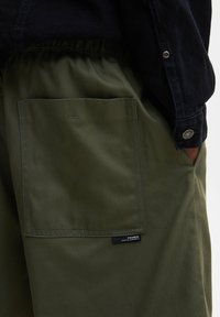 Gros plan sur un pantalon vert olive avec une grande poche carrée arrière et une taille élastique, main partiellement dans la poche, portant une manche de veste sombre.