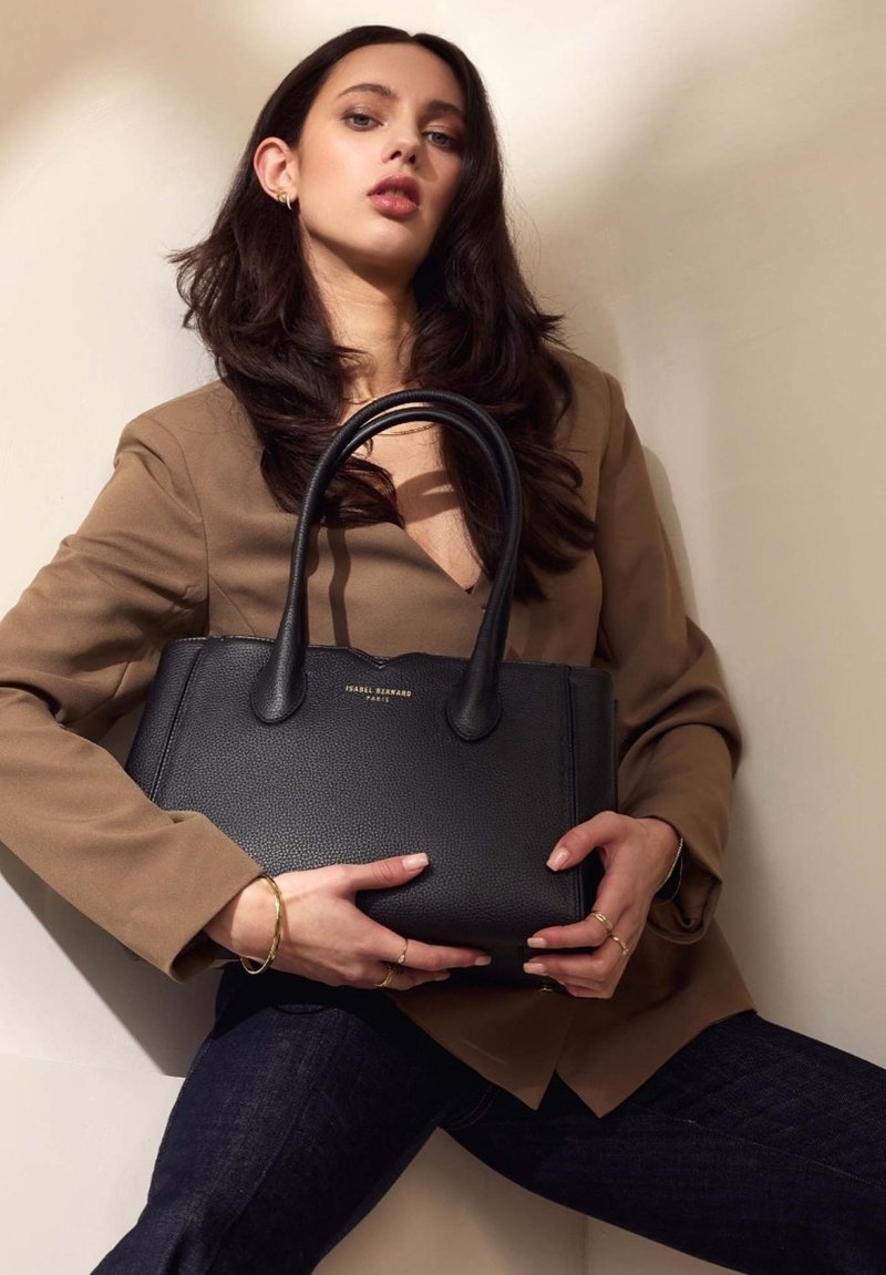 Schwarze Ledertasche mit doppelten Griffen, strukturierten Oberflächen und goldener Markenkennzeichnung. Das Modell trägt ein braunes Blazerjacke und dunkle Jeans, während es die Tasche hält.