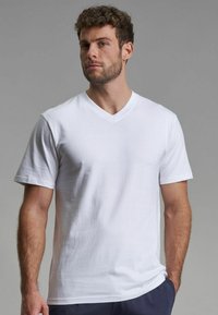 Weisses Baumwoll-V-Ausschnitt-T-Shirt mit kurzen Ärmeln, das eine glatte Textur und einen lässigen Schnitt aufweist, getragen von einem Mann vor einem neutralen Hintergrund.