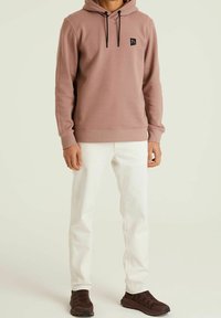 Felpa rosa con cappuccio, patch logo nero e cordini. Abbinata a jeans bianchi e scarpe sportive marroni. Texture liscia.